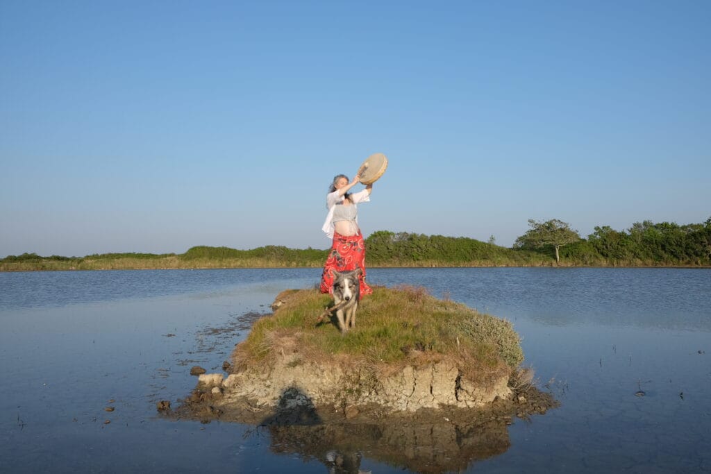 Photographie de Johanna Sandretto célébrant sa grossesse sur un ilôt entouré d'eau. Jouant du tambourin